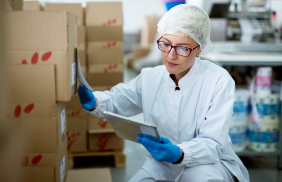 Young beautiful focused female worker in sterile cloths using a tablet to check correction of inventory in factory storage room.